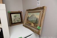 Two framed pictures: one oil painting of a mountain landscape in a gold frame, one floral picture tile in an ornate frame