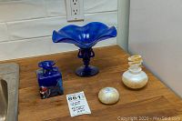 Blue glass pedestal bowl, blue square perfume bottle with multicolor detail, white perfume bottle with gold leaf and matching paperweight on wood surface.