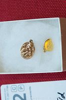 Two religious pendants side by side on white fabric inside a box, showing detailed engravings and gold tones.
