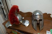 Two metal helmets displayed on wooden surface, one with red plume (Greek style) and one plain metal (Roman style).