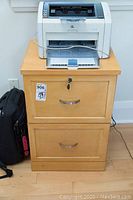 First wood-like 2-drawer filing cabinet with printer on top and black backpack nearby