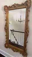 Full view of rectangular mirror with carved gilt wood frame hanging on wall, reflection of staircase and chandelier