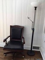 Vintage carved wood rolling desk chair with black leather seat and backrest, showing carved floral detail on upper backrest and wooden armrests.