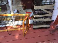 Two wood framed dog gates with metal grating placed on a deck beside a stack of folded, taped metal dog crates.