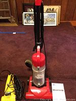 Dirt Devil red and gray upright vacuum cleaner showing power cord and clear dust collection canister, placed on a carpeted floor with picture frames in background.