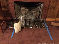 Wide shot of fireplace hearth with assorted items including old-fashioned glass milk bottles in metal carrier, two glass vases, large decorative floor vase with leaf design, and fireplace tools.