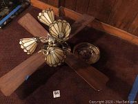 Ceiling fan viewed from an angle showing wooden blades and Tiffany-style lamps, on carpeted floor.