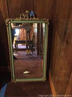 Full view of the rectangular mirror leaning against a wall, showing the carved wood frame with gilt overlay and light green inner border.