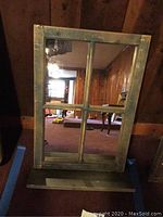Front view of green farmhouse style window pane mirror with shelf against wooden wall and carpeted floor.