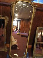 Full view of the tall mirror leaning against a wooden paneled wall, showing carved gilt frame with crest and etched glass floral designs.