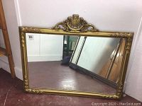 Full view of rectangular mirror on floor showing frame with gilt carved decoration and central crest at top.