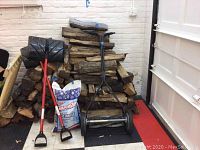 Stack of firewood logs with two red-handled snow shovels, manual grass cutter, and unopened ice melt bag arranged in front.