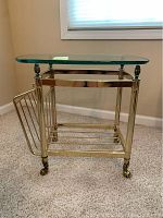 Side view of the mid century brass and glass side table, showing the thick curved glass top and brass frame with caster wheels and side magazine rack.