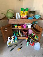 Full view of rolling wooden potting bench cabinet with numerous gardening tools, pots, gloves, sprays, and potting mix displayed on and around it.