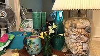 Photo showing shell lamp, decorative blue-green glass bottle, ceramic pitcher with flower design, green candles, and part of fan in background