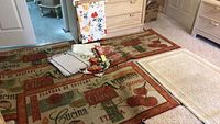 Four Tuscan print throw rugs and one cream colored throw rug on carpeted floor with assorted tablecloths and place mats laid on top of the rugs.