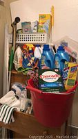 Photo showing assortment of cleaning and laundry products gathered in a red bucket with some items behind it on a wooden drawer, including detergents, sprays, and cleaning cloths