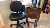 Black faux leather office chair shown with adjustable height and caster wheels, positioned beside two wooden folding tray tables.