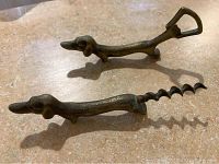 Metal dachshund bottle opener and corkscrew on countertop, both showing elongated dachshund shapes with a handle and corkscrew spiral.