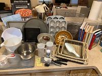Photo showing variety of baking pans including springform, muffin pan, bundt pan; mixing bowls; measuring cups and jugs; utensils including spatulas and basting brushes; bakeware racks and plastic lemon squeezer; several cookbooks on baking and cooking.