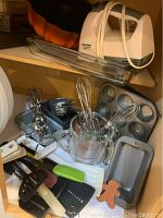 Photo showing multiple baking pans, electric hand mixer, utensils and measuring cups inside a cabinet shelf