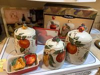 Overall kitchen-themed set including LeCreuset baking pan, three Venetian canisters, two fruit-shaped dishes, and the cutting board with place mat behind