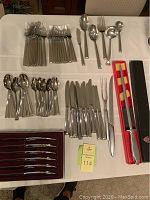 Full overview of the flatware assortment laid out on a white tablecloth, showing forks, spoons, knives, serving utensils, and boxed steak knives.
