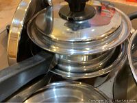Overhead close-up photo of stacked stainless steel pots and pans with lids and black handles.