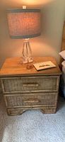 Nightstand with wicker rattan drawer, light brown color, topped with a table lamp and other small items, set on carpeted floor.