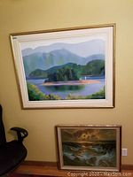 Larger painting of island and lake with framed gold border above smaller ocean wave painting, with a black chair nearby and beige wall background.