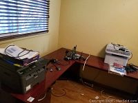 Samsung photocopier/printer on the left corner of a wooden desk, with cords on top, next to cordless phone handsets and base stations in the middle, and Brother printer on the right on another desk corner with some packing materials on top.