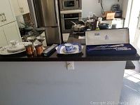 Wide shot showing carving set box with Birks Regency plating set on blue satin lining, alongside Royal Albert bone china cup, saucer and plate, plus other tableware items on kitchen island.