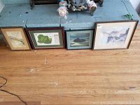 Four framed artworks placed on a wooden floor, from left to right: a small watercolor, Augusta golf map, landscape painting, and largest warped watercolor with stains