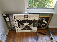 Photo showing three original mixed media art pieces leaning against a wall beneath a window. Pieces measure 24x24 each. One piece displays a black and white motif with 'Manhattan New York' text and a clock image; another has white bird figures on a dark, textured background; the third has a dark abstract design.