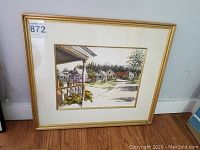 Full view of original village scene artwork in a gold frame, leaning against a white wall on wooden floor.