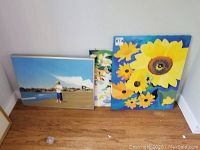 Two framed paintings resting against a wall on wooden floor, one floral artwork with yellow sunflowers and one beach scene with child.