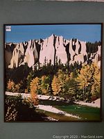 Full view of the framed square mountain scene painting showing rocky white peaks, autumn colored trees, and green river underneath a blue sky.