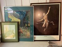 Three framed art pieces leaning against a wall: a botanical print with green mat, a 'Triumph' nude figure print, and a Starry Night style urban night scene.