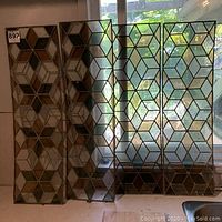 Four stained glass panels displayed indoors, arranged side by side blocking light from a window behind. Each panel has a repeating geometric cube/star pattern in shades of white, green, amber, and clear textured glass framed by metal cames.