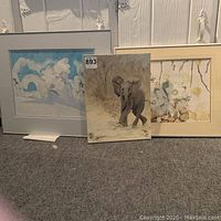 Three original artworks displayed together on floor: an elephant painting and two framed watercolors, one with floral motif and one with children on a swing.