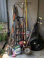 Group of assorted garden tools including hand tools with wooden handles, a hand roller, gardening supplies like coated lime and oil container, and the Mantis rototiller placed on the floor under a shelter.