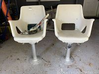 Two white boat chairs each mounted on metal pedestal bases placed indoors on a concrete floor.