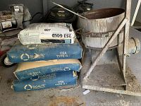 Photo of the cement mixer and four bags of cement mix stacked next to it. The bags include three Basalite Type GU and one 253 Gold. Mixer is metal with signs of cement residue.