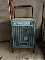 Full front view of blue Drizair 110 dehumidifier on floor against wall, showing handle and legs.