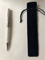 Silver pen with transparent midsection containing white Swarovski crystals and etched silver top barrel, next to black drawstring fabric case.