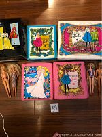 Five vintage Barbie doll carrying cases with colorful illustrated covers placed on floor alongside five vintage Barbie dolls in various states of dress.