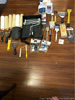 Overview of painting supplies lot spread on wooden floor including rollers, scrapers, brushes, trays, and bottles