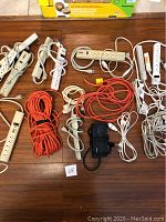 Collection of various extension cords and power strips laid on a wooden floor, including white power strips with switches and orange heavy-duty cords.