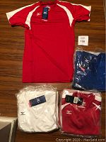 Photo showing a red Mizuno volleyball shirt laid flat, with two packaged red shirts, one white shirt, and one packaged blue shirt visible. All shirts have tags attached.