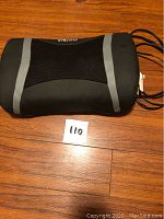Front view of HoMedics shiatsu lumbar massage pillow showing mesh black center with gray sides, placed on wooden floor with lot number card.